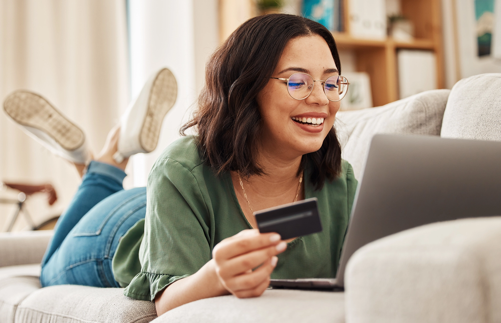 Woman smiles while entering her card information into her computer to purchase an item while lying on the couch